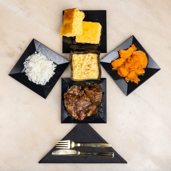 A square plate of meat with gravy, surrounded by sides of rice, mac and cheese, candied yams, and cornbread.