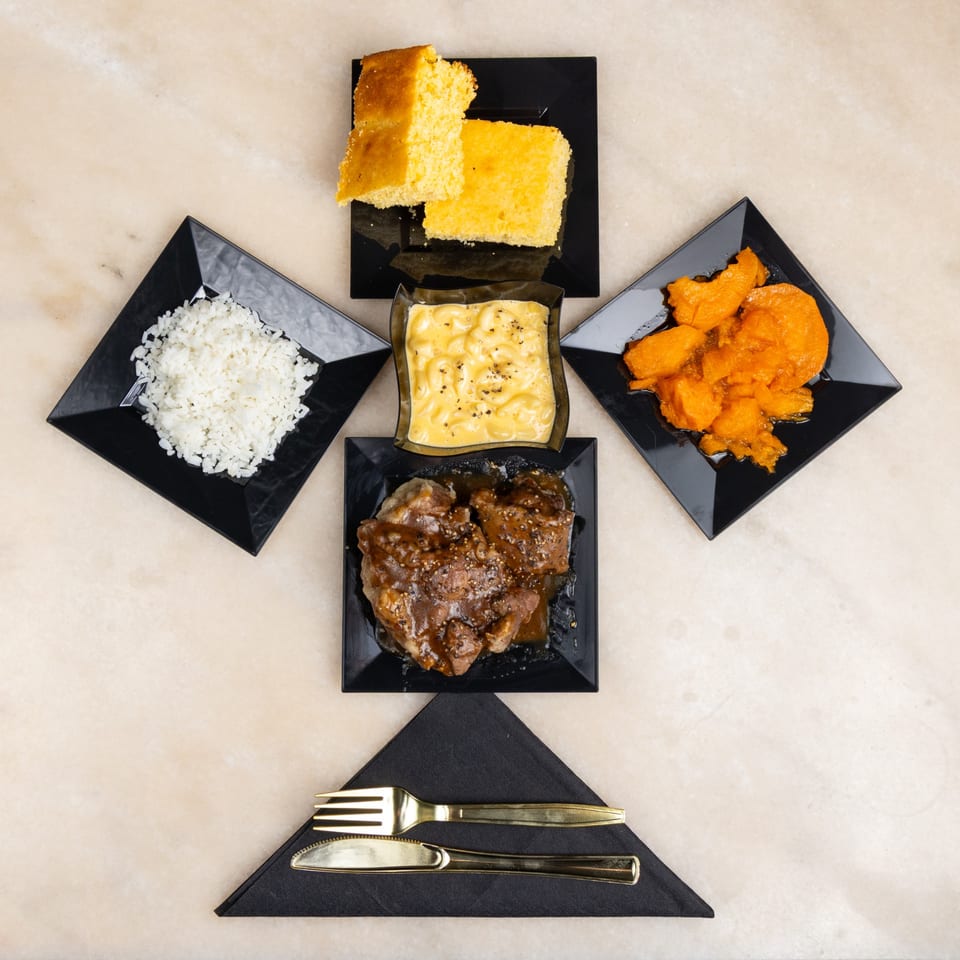 A square plate of meat with gravy, surrounded by sides of rice, mac and cheese, candied yams, and cornbread.