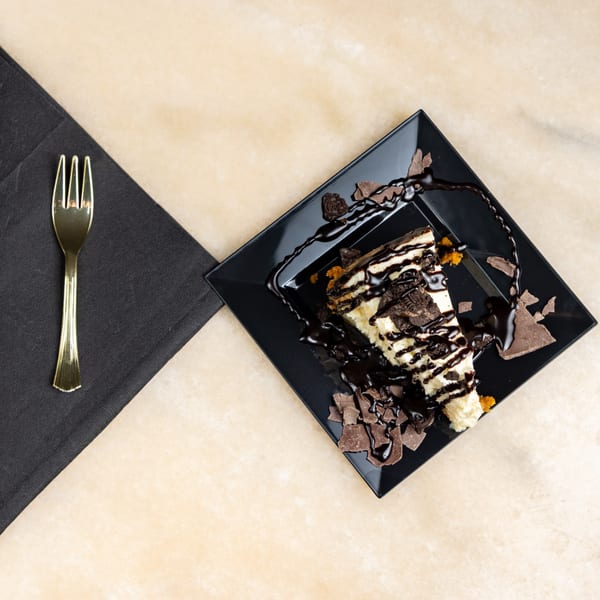 A slice of cheesecake with chocolate syrup, cookie crumbles, and chocolate shavings on a black plate.