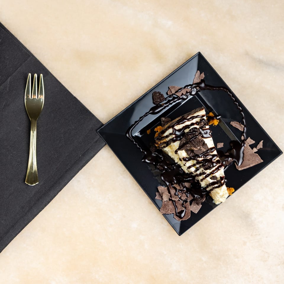 A slice of cheesecake with chocolate syrup, cookie crumbles, and chocolate shavings on a black plate.