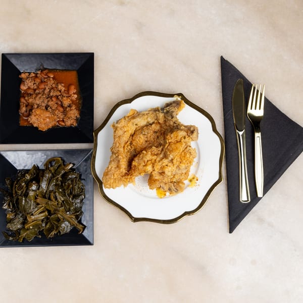 Fried chicken, collard greens, and baked beans on black and white plates with gold cutlery.