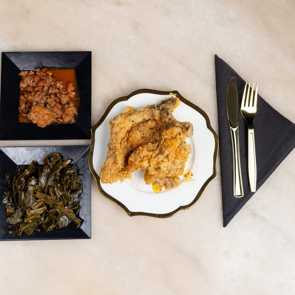 Fried chicken, collard greens, and baked beans on black and white plates with gold cutlery.