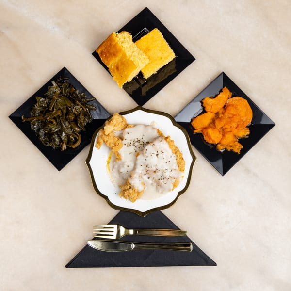 Fried chicken with gravy, collard greens, sweet potatoes, and cornbread on black and white plates.