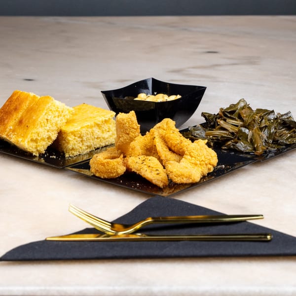 Southern meal with fried fish, collard greens, mac and cheese, and cornbread on black plates.