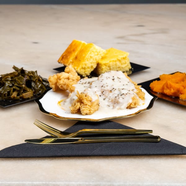 Fried chicken with gravy, collard greens, sweet potatoes, and cornbread on a table with gold cutlery.