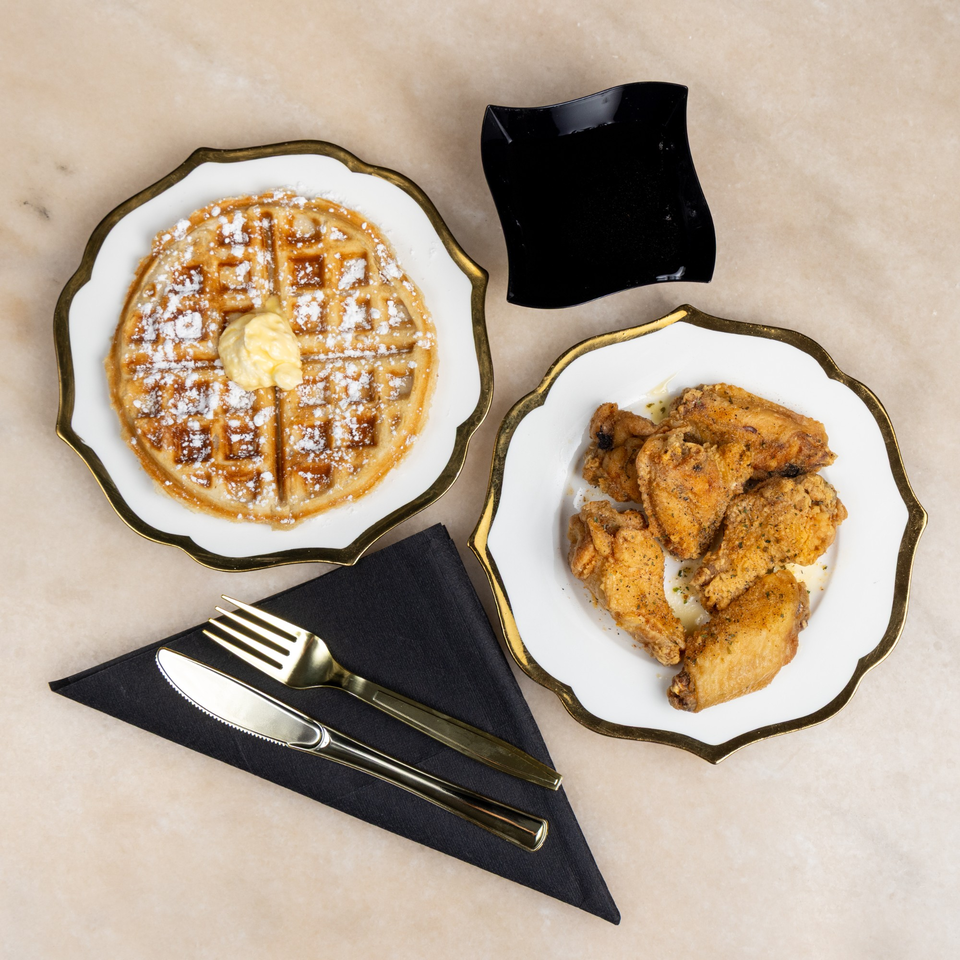 Golden waffle with butter and powdered sugar, fried chicken wings, and black syrup dish on a marble surface.